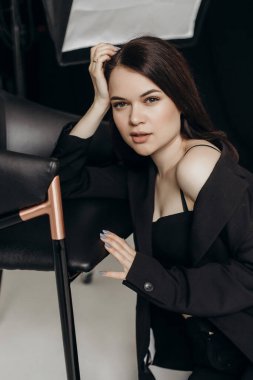 A girl in a black suit on a white background sits on the floor near a chair