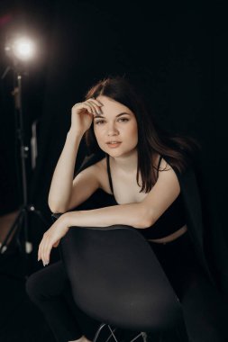 A girl in a black suit on a black background sits on a chair