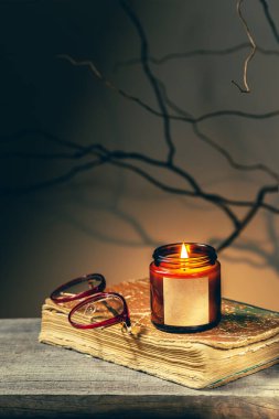 Eski bir antika kitap, gece yanan cam bir kavanozda gözlük ve mum. Ağaç brunch 'ları ve gölgeler ürkütücü karanlık akademi atmosferini oluşturur. Sıcak sonbahar okuma zamanı