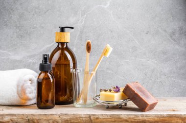 Beyaz havluyla banyo masası, sıvı cilt bakım ürünleriyle dolu koyu cam şişeler, bambu diş fırçaları ve gri mermer arka planda el yapımı katı sabunlar.