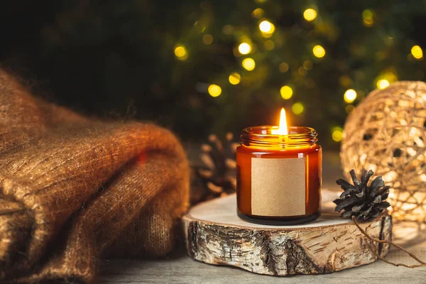 Cozy winter card with burning aroma candle in a dark glass jar with empty label mock up and wool sweater over blurred bokeh lights on the background. Christmas holiday at home.