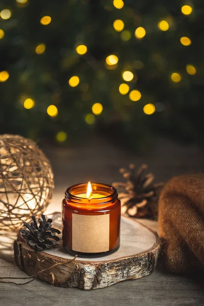 Cozy winter evening card with burning aroma candle in a dark glass jar with empty label mock up. Soft focus, copy space for text. Blurred bokeh lights on the background. Christmas holiday at home.