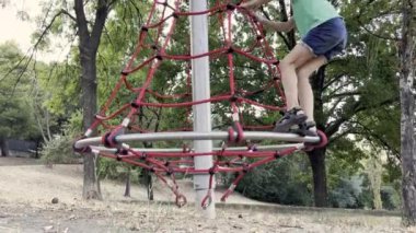 7 yaşında beyaz bir çocuk parktaki çocuk parkında ağa tırmanıyor. Çocuk halatla tırmanırken eğleniyor.