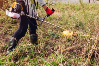 Bahçıvan çayırda çalı biçme makinesiyle çim biçiyor. Metal bıçak diski ile manuel benzin budayıcı ile kuru çimleri buduyor. Çizme giyen çiftçi.