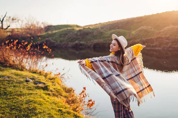 Šťastná žena odpočívající u podzimního jezera při západu slunce. Stylová dívka v klobouku hraje s větrem a šála těší podzim krajiny — Stock fotografie