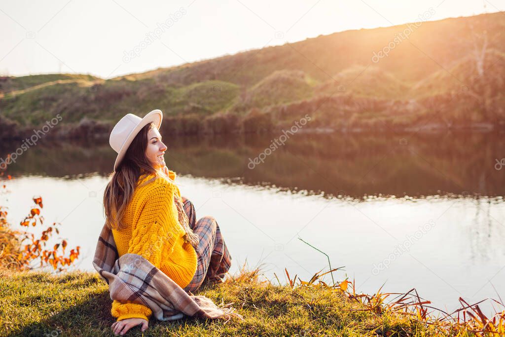 Young woman relaxing by autumn lake at sunset. Stylish girl in hat sitting on bank enjoying fall landscape, breathing freely. Mental health. Healing nature