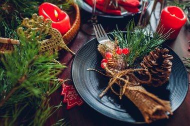 Köknar dalları, kırmızı mumlar, koniler ve süslemelerle süslenmiş iki kişilik Noel yemeği masası. Altın geyiklerle çevrili çatal bıçaklı ve gözlüklü tabaklar.