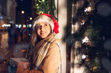 Noel Baba şapkalı mutlu kadın, Noel 'de şehir caddesinde mağaza vitrininin hediye kutularını taşıyor..