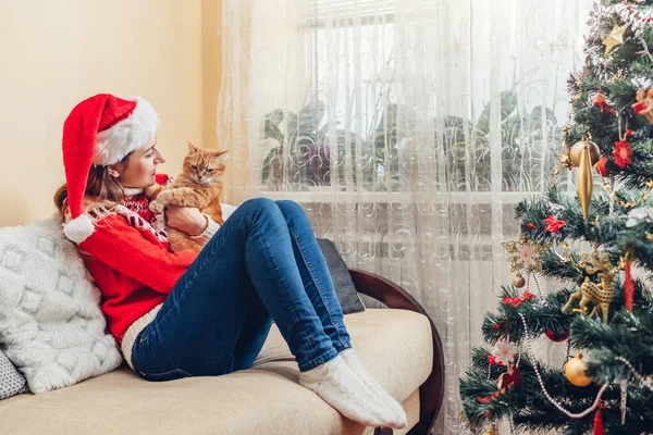 Boże Narodzenie z kotem. Kobieta gra i przytula zwierzaka w kapeluszu Mikołaja przez Nowy Rok drzewa w domu. — Zdjęcie stockowe