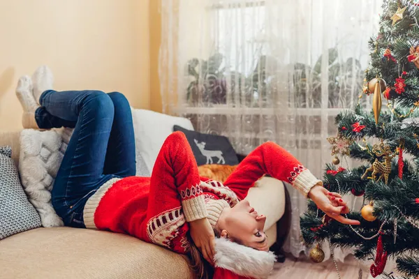 Znudzona, rozczarowana młoda kobieta leżąca na kanapie przy choince w kapeluszu Mikołaja smutna, zdenerwowana, samotna — Zdjęcie stockowe