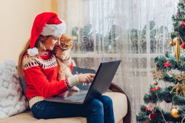 Kediyle Noel kutlaması. Noel Baba 'nın şapkalı kadın evdeki yılbaşı ağacının yanında dizüstü bilgisayar kullanıyor. İzolasyonda internet kullanarak tatilde aile rahatlıyor