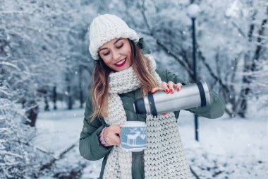 Karlı kış parkında vakum tüpünden sıcak çay döken bir kadın. Gri örülü Noel süveteri ya da geyikli palto giymiş, onu sıcak tutacak bir bardak.