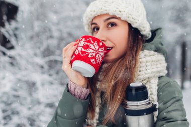 Karlı kış parkında elinde elektrikli süpürge şişesiyle çay içen kadın manzaranın tadını çıkartıyor. Kırmızı örülmüş bir kupa Noel süveteri