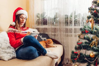 2022 yılının gol listesi yazan genç bir kadın. Yılbaşı ağacının yanında oturuyor. Noel Baba şapkası ve kalın süveter giyiyor..