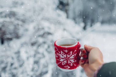Kış parkında kırmızı örülmüş Noel ceketi giymiş bir fincan çay ya da kahveyi kapat. Soğuk ve karlı havada ısınmak için içecekler.