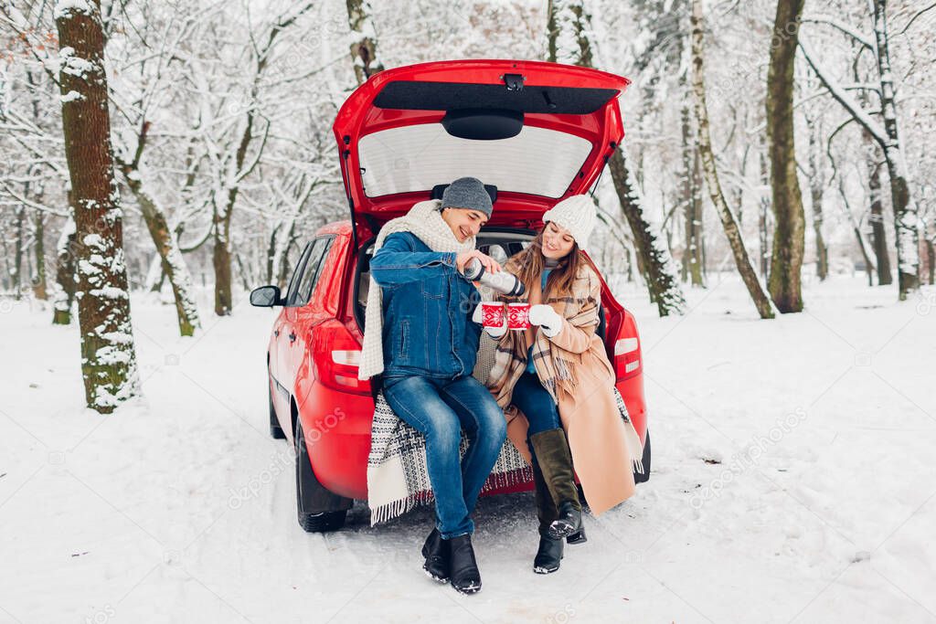 Couple in love sitting in car trunk having hot tea in snowy winter forest. Man pouring drink from thermos. People relaxing outdoors during road trip