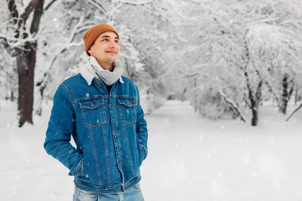 穿着牛仔裤外套帽走在户外的雪地冬季公园里的时尚男子的画像 英俊的小伙子喜欢下着雪 欣赏风景 男性时尚 — 图库照片