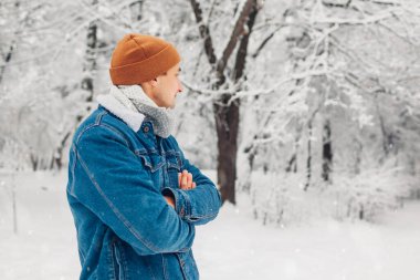 Karlı kış parkında, elleri açık havada kesişen kot şapkalı şık bir adamın portresi. Yakışıklı adam kar ve manzara yağdırmayı seviyor. Erkek modası