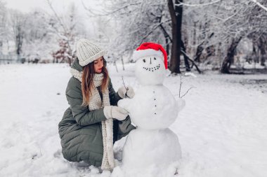 Kız, kardan adamı açık havada, karlı kış parkında Noel Baba şapkası, atkı ve dallarla süslüyor. Noel eğlence aktiviteleri