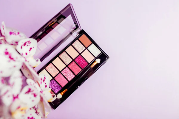 Palette of new colourful eyeshadows, pigments, glitter with orchid flowers. Make-up products on purple background — Stockfoto