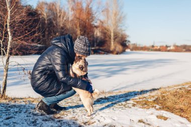 Woman walking pug dog in snowy winter park by frozen lake hugging pet. Happy puppy wearing harness standing on hind legs.