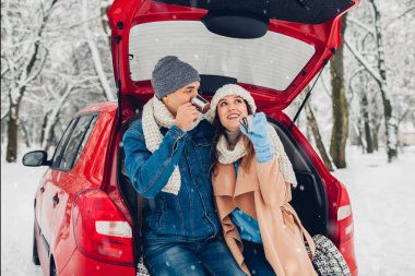Valentine's day. Couple in love sitting in car trunk drinking hot tea in snowy winter forest and chatting. People relaxing outdoors during road trip