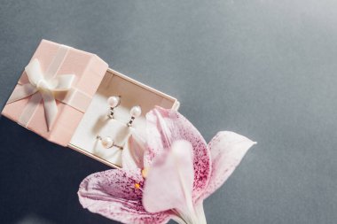 Sevgililer günü hediyesi. Çiçekli pembe hediye kutusunda bir inci mücevher seti. Küpe ve yüzük ve gri arka planda zambak. Boşluğu kopyala