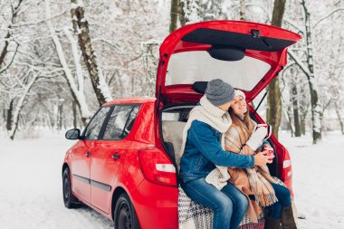 Sevgililer Günü aşığı çift arabanın bagajında oturmuş karlı kış ormanlarında sıcak çay içiyor. İnsanlar romantik bir buluşmada tatillerini kutlarken rahatlıyorlar.