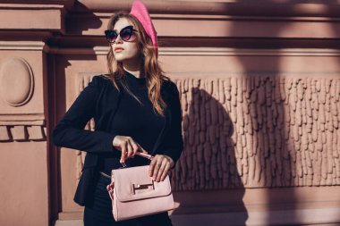 Kadın güzelliği, moda. Pembe bereli şık bir kadın, şehir caddesinde gözlüklü çanta taşıyor. Bahar giysileri, aksesuarlar.