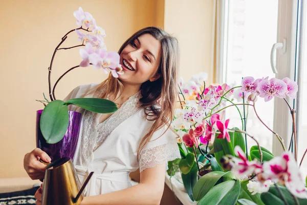 Улыбающаяся женщина восхищается цветущей фиолетовой орхидеей, держащей горшок. Девушка садовница заботится о домашних растениях и цветах. — стоковое фото