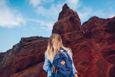 Yunanistan 'ın Santorini adasındaki Akrotiri plajında yürüyen bir turist. Sırt çantalı kadın yürüyüşçü yüksek kayalara bakarak manzaranın tadını çıkarıyor. Düşük açı