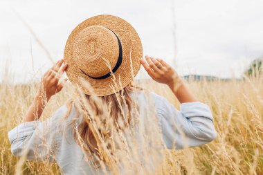 Female fashion. Back view of young woman walking among high grasses in summer meadow wearing straw hat and linen shirt. Stylish accessories and clothing