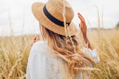 Back view of young woman walking among high grasses in summer meadow wearing straw hat and linen shirt. Harmony and balance