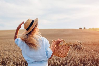 Back view of young stylish woman in hat walking in summer field holding straw handbag with wheat bundle at sunset. Harmony and freedom