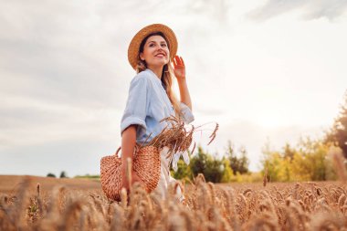 Portrait of happy young woman walking among wheat in summer field wearing straw hat holding handbag at sunset. Girl relaxing enjoying landscape
