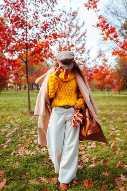 Full body portrait of young woman wearing stylish warm clothes in autumn park among red trees. Female fall fashion. Modern accessories