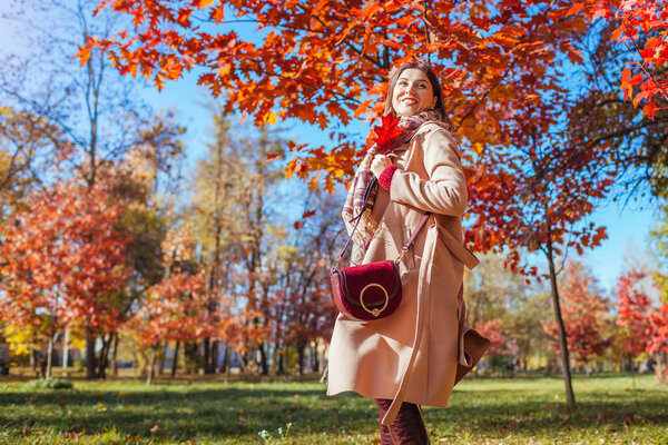 Happy young woman walking in autumn park among red trees. Female fall fashion. Stylish girl wearing beige coat and holding purse