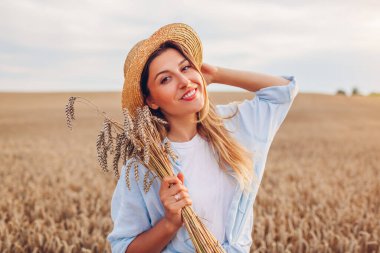 Portrait of happy young woman walking among wheat in summer field wearing straw hat holding bundle of ripe wheat at sunset. Rustic boho style