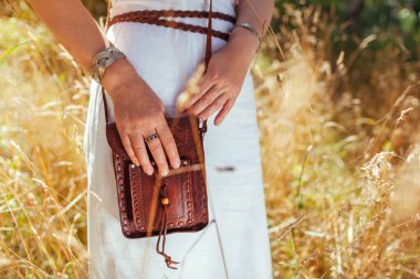Close up of brown boho leather purse. Stylish woman wearing white dress, jewelry, holding handbag outdoors. Fashionable female accessories