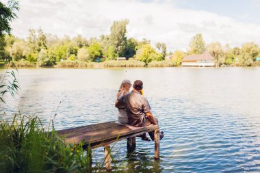 Senior family couple relaxing by summer lake. Man and woman enjoying natural landscape and hugging sitting on pier