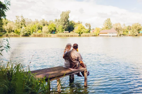 Senior family couple relaxing by summer lake. Man and woman enjoying natural landscape and hugging sitting on pier