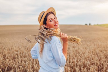 Portrait of happy young woman walking among wheat in summer field wearing straw hat holding bundle of ripe wheat at sunset. Rustic boho style