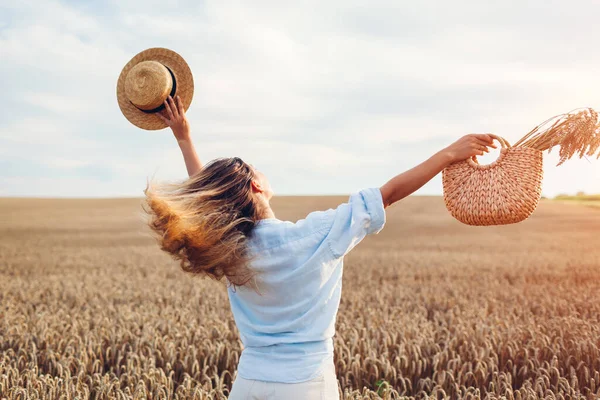 Back view of young happy woman jumping enjoying view in summer field holding straw handbag with wheat bundle at sunset. Harmony and freedom