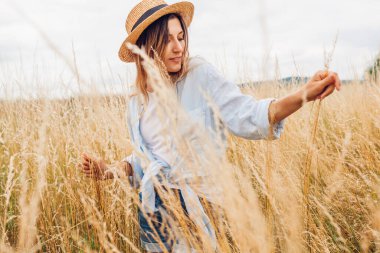 Portrait of young woman walking among high grasses in summer meadow wearing straw hat and linen shirt enjoying nature. Harmony and balance