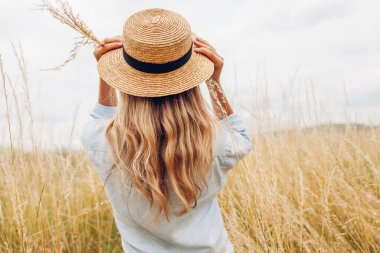 Female fashion. Back view of young woman walking among high grasses in summer meadow wearing straw hat and linen shirt. Stylish accessories and clothing. Space