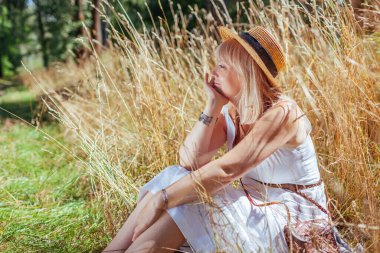 Stylish middle-aged woman relaxing sitting in summer meadow surrounded with dry high grass outdoors. Harmony with nature