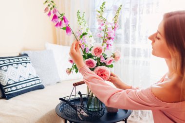 Woman makes bouquet of roses and foxgloves flowers in vase on table at home. Interior and summer decor. Fresh pink orange blooms of austin and digitalis in arrangement