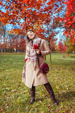 Güz parkında kırmızı ağaçların arasında şık, kalın kıyafetler, botlar ve aksesuarlar giyen güzel bir kadının vücut portresi. Kadın sonbahar modası.