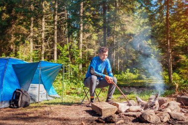 Ormanda kamp ateşinde dinlenen bir gezgin çadırın yanında oturmuş yangını kontrol ediyor. Yaz kampı. Yalnız seyahat etmek, doğanın tadını çıkarmak