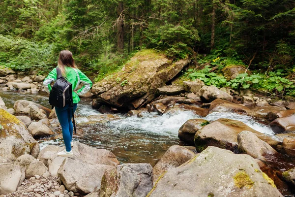 Stock image Back view of female traveler hiker with backpack admiring landscape by mountain river in Carpathian forest. Active sportive lifestyle